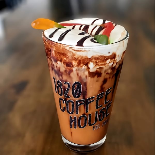 A tall glass of iced coffee with whipped cream, chocolate drizzle, and gummy worms on a wooden table.