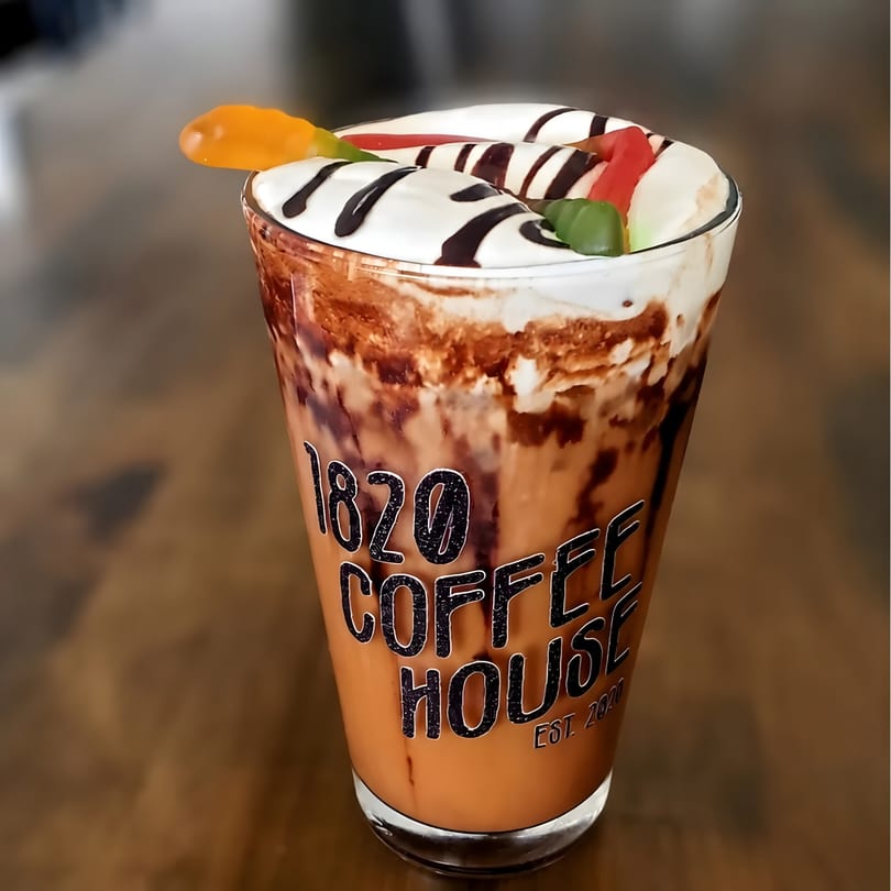 A tall glass of iced coffee with whipped cream, chocolate drizzle, and gummy worms on a wooden table.