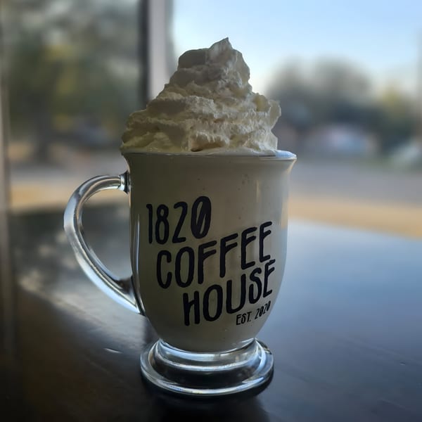 A clear mug from 1820 Coffee House filled with a drink and topped with whipped cream.