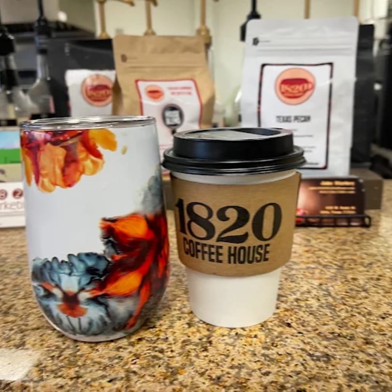 A coffee cup from 1820 Coffee House and a colorful tumbler sit on a counter with coffee bags.
