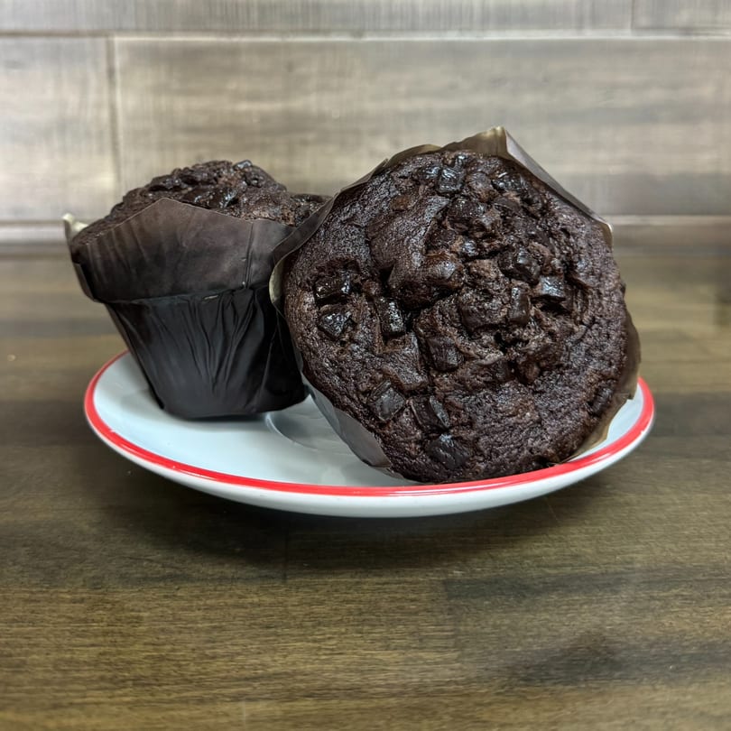 Two dark chocolate muffins with chocolate chips in brown wrappers on a white plate with a red rim.