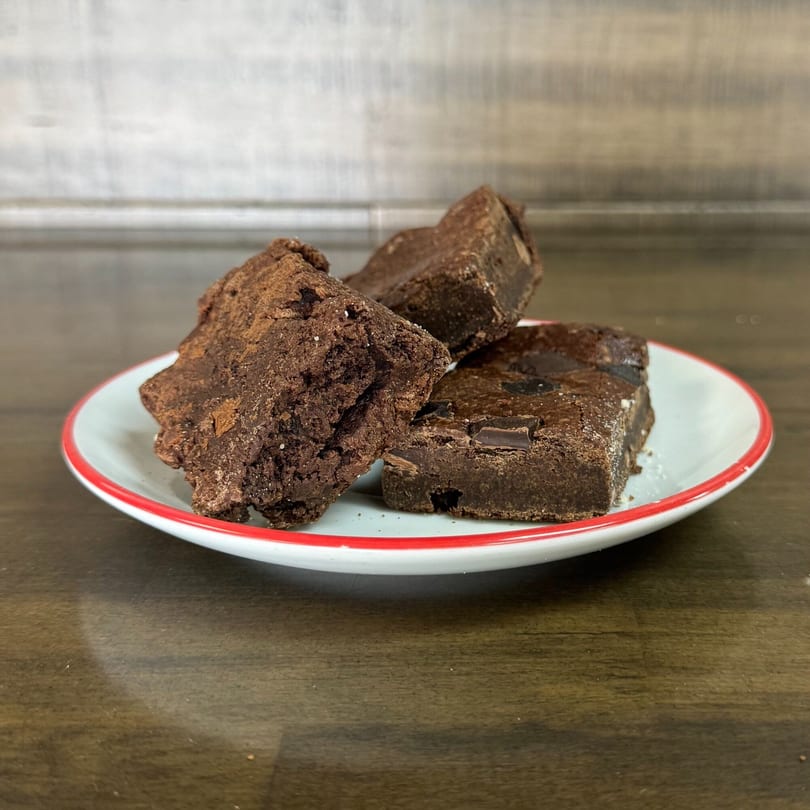 Three rich chocolate brownies, one with chocolate chips, on a white plate with a red rim.