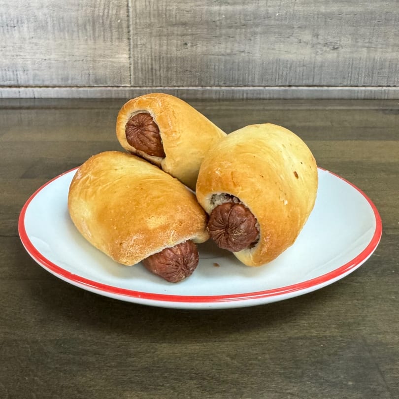 Three golden-brown pigs in a blanket, with hot dogs peeking out, on a white plate with a red rim.